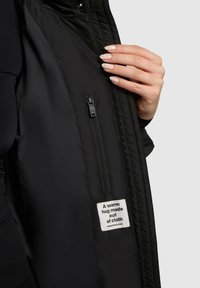 Sort isoleret jakke med en lynlåslomme og et stofmærke, der siger "Et varmt kram lavet af stof." Glat tekstur og strømlinet design.