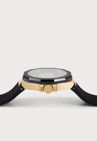 Orologio ottagonale nero e oro con una lunetta texturizzata, quadrante in vetro trasparente e cinturino in gomma. Presenta una corona centrale di lato.