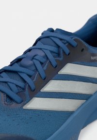 Μπλε αθλητικό παπούτσι με υφασμάτινη υφή και συνθετικά επικαλύμματα, με τρεις αντανακλαστικές ασημί ρίγες και κορδόνια στο ίδιο μπλε χρώμα.