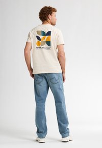 Krémové tričko s barevným geometrickým designem na zádech, kombinované se světle modrými denimovými džíny a bílými teniskami.
