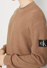 Brązowy sweter dzianinowy z prążkowanymi mankietami i czarną łatą z logo na lewym ramieniu. Posiada okrągły dekolt i teksturowany materiał.