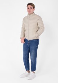 Giacca trapuntata beige con colletto alto, zip frontale e tasche laterali, abbinata a jeans azzurri chiari e sneakers bianche. Texture liscia.