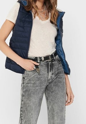 Vrouw met gelaagde zilveren kettingen, wit t-shirt, marineblauw gewatteerd vest, zwarte riem en grijze jeans met zuureffect, met hand in de zak.