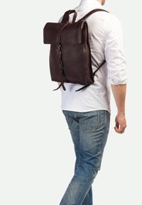 Mochila de cuero marrón con cierre de solapa y correas ajustables. Textura suave y diseño minimalista con un único detalle de hebilla en la parte frontal.