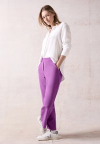 Chemise blanche texturée avec une poche, associée à un pantalon violet ajusté et des baskets blanches. Pose de standing, mains dans les poches. Fond neutre.