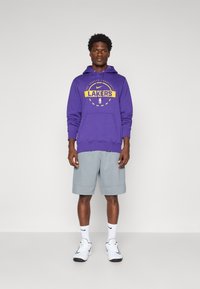 Purple hoodie with "LAKERS" graphic in yellow, paired with gray shorts. Model wears white sneakers and Nike logo socks.