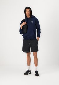 Marinegenser, svarte stripete shorts og svarte joggesko. Inkluderer en liten svart skulderveske. Minimalistisk design med kontrasterende detaljer.