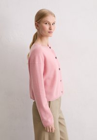 Gilet rose ajusté avec un col rond, fabriqué en tissu doux, doté de boutons marron sur le devant et de poignets côtelés.