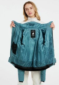Eine türkisfarbene Outdoor-Jacke mit einem durchgehenden Reißverschluss, mehreren Innentaschen und einer glatten Textur. Enthält Logo- und Markendetails im Inneren.