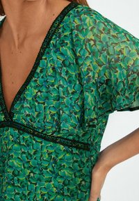 Blusa floreale verde con scollatura a V profonda, maniche a campana e dettagli in pizzo nero lungo il collo e l'orlo. Materiale leggero e trasparente.