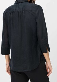 Chemise en lin noire avec boutons, pli dans le dos, manches trois-quarts, fentes latérales et ourlet arrondi. Texture lisse et coupe décontractée.