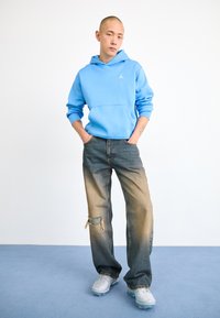 Sweatshirt à capuche bleu clair avec poche avant, associé à un jean en denim délavé avec des déchirures effilochées, et des baskets gris clair.