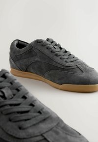 Adidași din suede gri, cu profil redus, având vârful rotunjit, talpa din cauciuc și un design clasic cu șireturi; textură netedă și detalii minime.