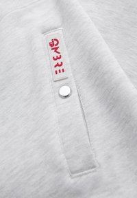 Gros plan sur un tissu gris clair avec une poche verticale dotée d'un bouton-pression argenté et une petite étiquette blanche avec le texte rouge "AM3RE".