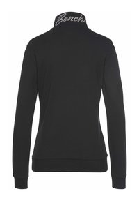 Black long-sleeve jacket with high collar featuring white cursive "Bench" logo embroidery, shown from the back on a white background.