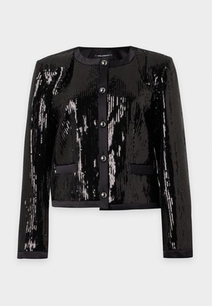 Femme portant une veste noire à sequins, un top blanc à col montant, un pantalon noir, des sandales à talons noires et tenant un petit sac à main noir clouté.