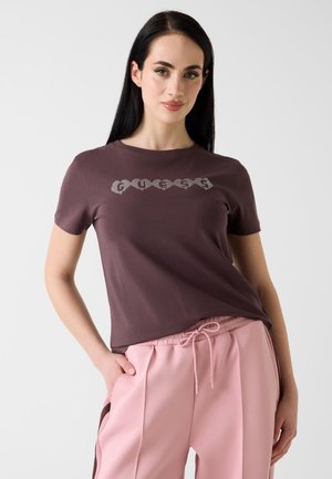 Femme aux longs cheveux noirs portant un t-shirt marron foncé à manches courtes et un pantalon rose clair à cordon, debout avec une main dans la poche.