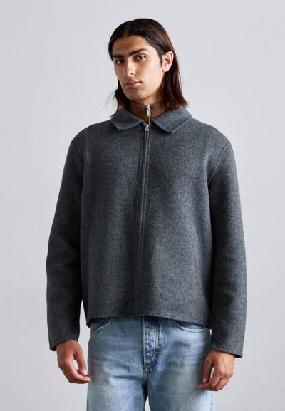 Veste en laine grise avec un col, fermeture éclair à l'avant et manches longues, offrant une coupe décontractée et une texture lisse. Portée sur un jean en denim.