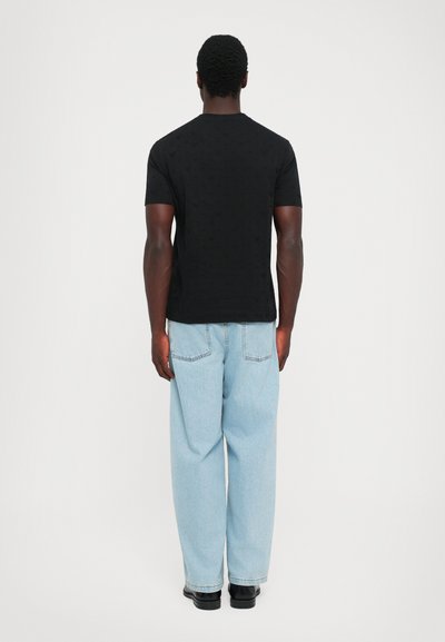 Mann, der mit dem Rücken zur Kamera steht, trägt ein schwarzes Kurzarmhemd, helle, lockere Jeans und schwarze Schuhe vor einem schlichten weißen Hintergrund.