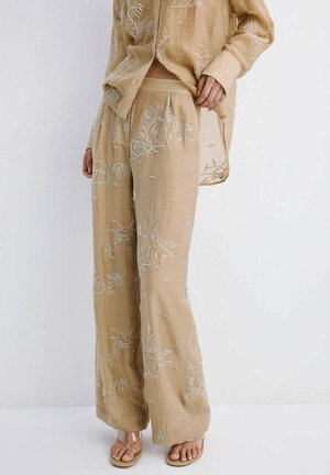 Pantalones beige holgados con bordado floral blanco, combinados con una camiseta de manga larga a juego y sandalias beige abiertas en la puntera sobre fondo blanco.