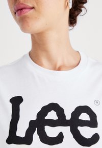 Vit bomulls-T-shirt med en stor svart logotyp som står "Lee." Närbild av halslinningen och en del av ansiktet; slät textur är tydlig.