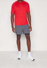 Červené sportovní tričko s krátkým rukávem a černým logem, spárované se šedými sportovními šortkami s černým akcentem a bílými teniskami.
