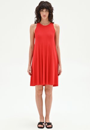 Donna con capelli ricci e scuri che indossa un vestito rosso senza maniche, plissettato e di lunghezza al ginocchio, e sandali neri piatti, in piedi su uno sfondo bianco.