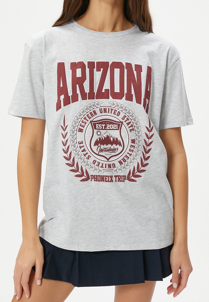 Femme portant un T-shirt gris avec un texte bordeaux indiquant "ARIZONA" et "Western United State Phoenix Trip" avec un graphique de montagne au centre.