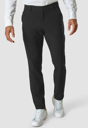 Homme portant un pantalon noir slim, une chemise blanche et des baskets blanches, debout devant un fond uni.