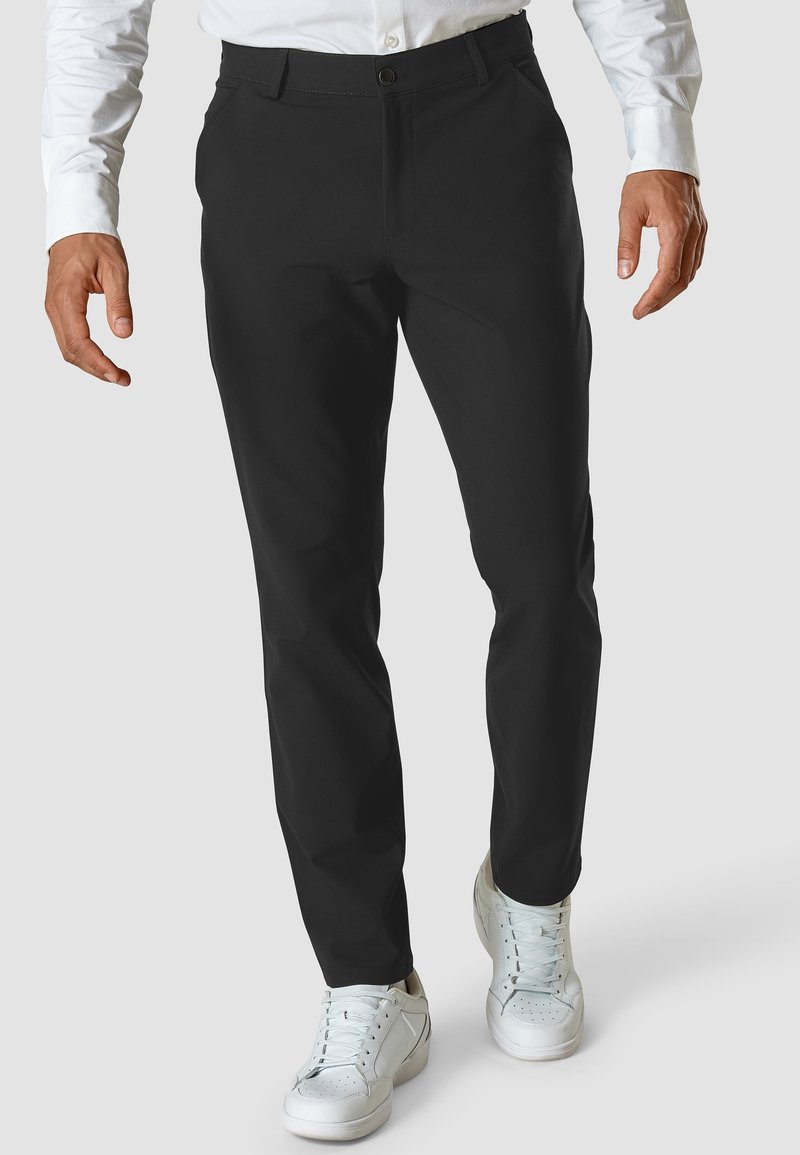 Homme portant un pantalon noir slim, une chemise blanche et des baskets blanches, debout devant un fond uni.