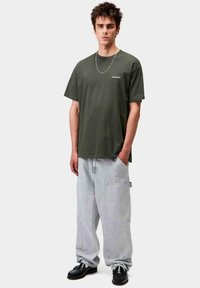 Joven de pie, vistiendo una camiseta Carhartt verde oscuro, pantalones anchos gris claro, zapatos negros y un collar delgado de cadena plateada.