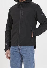 Mężczyzna w czarnej, zapinanej na zamek, kapturowej kurtce z zamkiem na piersi i kieszeniach, regulujący pasek rękawa, w połączeniu z niebieskimi dżinsami.