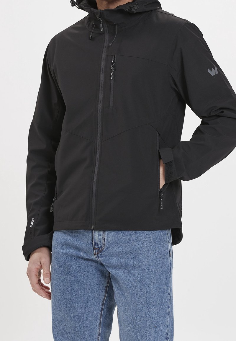 Mężczyzna w czarnej, zapinanej na zamek, kapturowej kurtce z zamkiem na piersi i kieszeniach, regulujący pasek rękawa, w połączeniu z niebieskimi dżinsami.