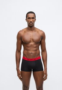 Μαύρα μπόξερ σορτς με αντίθετη κόκκινη μέση που φέρει το λογότυπο της μάρκας. Λείο ύφασμα με εφαρμοστή σχεδίαση.