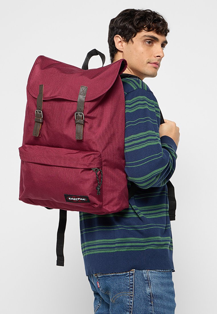 Junger Mann, der einen navy- und grün gestreiften Pullover und Jeans trägt, mit einem großen burgunderfarbenen Eastpak-Rucksack auf dem Rücken.