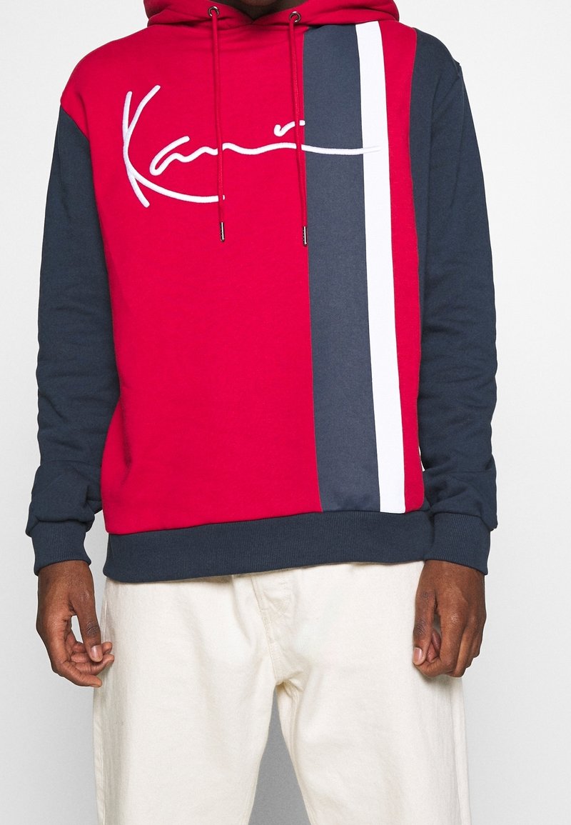 Hombre vestido con una sudadera roja y azul marino con rayas verticales blancas y azul marino y un texto en blanco, combinada con pantalones color crema.