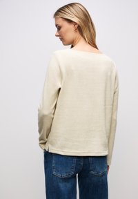 Beiger Strickpullover mit weitem Halsausschnitt, langen Ärmeln und seitlichen Schlitzen. Kombiniert mit blauen Jeans. Die Textur wirkt weich und lässig.