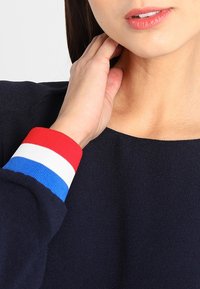 Navy-blaues Oberteil mit rundem Halsausschnitt und langen Ärmeln, das an den Bündchen in Rot, Weiß und Blau gestreift ist. Glatte Textur.