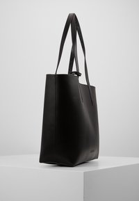 Czarna skórzana torba tote o eleganckim wzornictwie, gładkiej teksturze, długim pasku i minimalistycznym logo blisko podstawy.