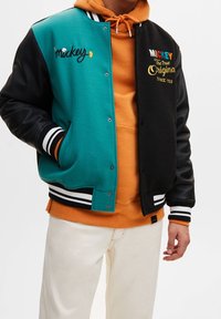 Veste universitaire pour hommes, devant en laine bleu sarcelle, manches en cuir noir, poignets côtelés à rayures, logo brodé "Mickey", portée par-dessus un sweat à capuche orange.