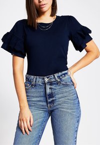 Marinblå topp med korta puffärmar och spetskant, kombinerad med höga blå denimjeans. Enkel rund halsringning och figursydd design.