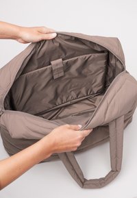 Ręce trzymają otwartą taupe'ową torbę z wyściółką, ukazując dużą komorę na zamek błyskawiczny z wyściełanym miejscem na laptopa w środku.