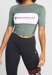 Top deportivo corto verde con banda blanca que dice "TOMMY SPORT", combinado con leggings negros de cintura alta que exhiben un logotipo rojo y blanco.