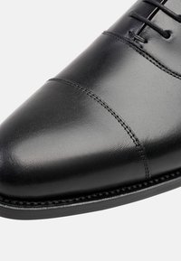 Chaussure de ville en cuir noir avec un bout pointu, des coutures décoratives, une texture lisse et une semelle en caoutchouc noir fine. Conçue pour les occasions formelles.