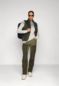 Chaleco acolchado verde sobre un suéter estampado, pantalones verde oliva y zapatos blancos. Los accesorios incluyen una mochila negra y gafas de sol.