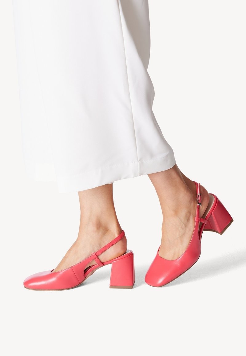 Pieds chaussés de chaussures à talons blocs corail rose à bride arrière, associés à un pantalon blanc à jambes larges sur un fond uni.