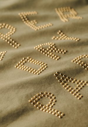 Tissu vert olive avec des clous dorés surélevés formant des lettres et des formes, créant un motif texturé sur la surface.