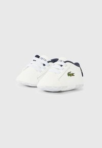 Λευκά αθλητικά παπούτσια με ομαλή υφή, διαθέτοντας ναυτικό μπλε λεπτομέρειες και ελαστικά κορδόνια. Κεντημένο πράσινο λογότυπο κροκοδείλου στην εξωτερική πλευρά.