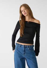 Crna majica dugih rukava s naboranom teksturom, off-the-shoulder dizajnom i rastezljivim materijalom, u kombinaciji s trapericama visokog struka plave boje.