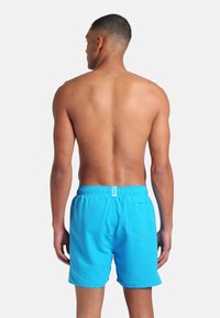 Hombre con cabello corto de pie de espaldas, vistiendo pantalones de baño azul brillante con un bolsillo trasero, contra un fondo blanco liso.